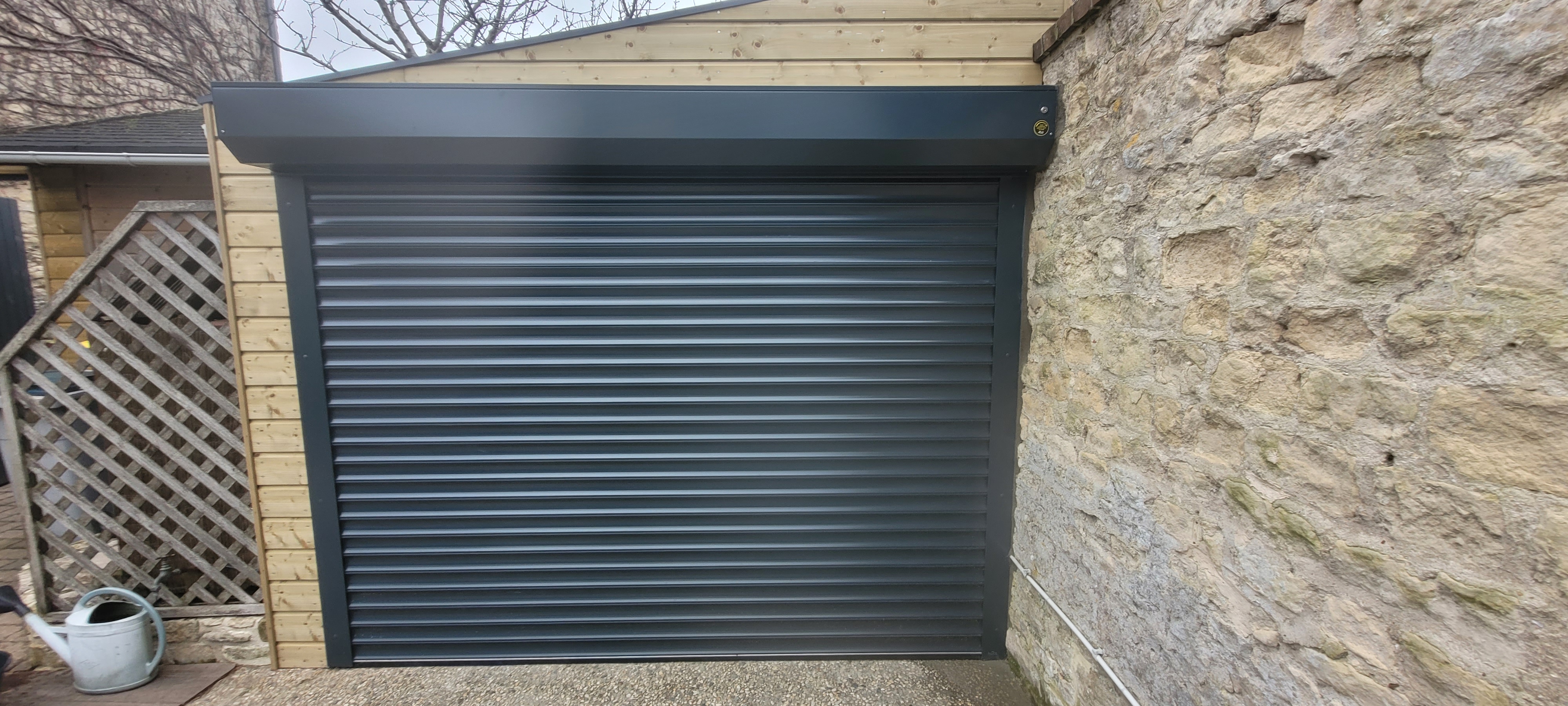 Porte de garage roulante noire et mur en pierre.