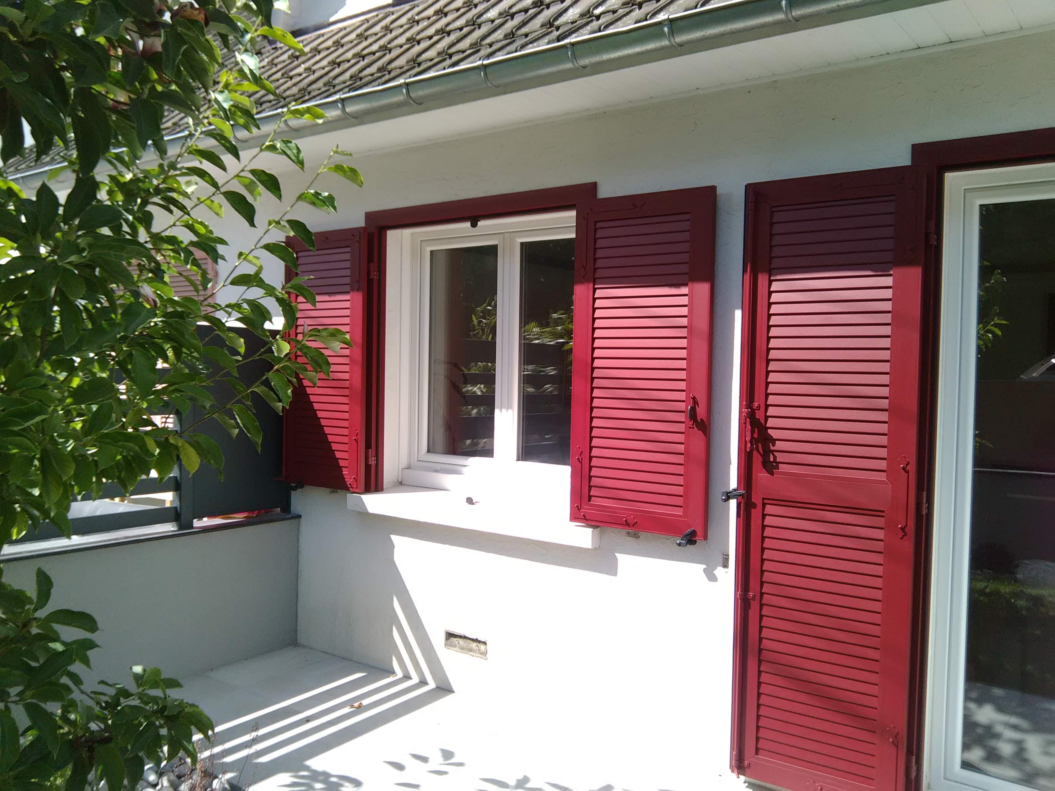 Fenêtre avec volets rouges ouverts sur maison blanche.